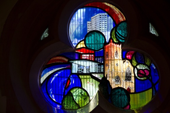 Close up of a contemporary stained glass window at St. John's Church, Ladywood. With the high-rises in the background, trees and shrubs in the foreground at the church itself in the middle, it symbolises that St. John's stands at the heart of the inner-city community that it serves.
